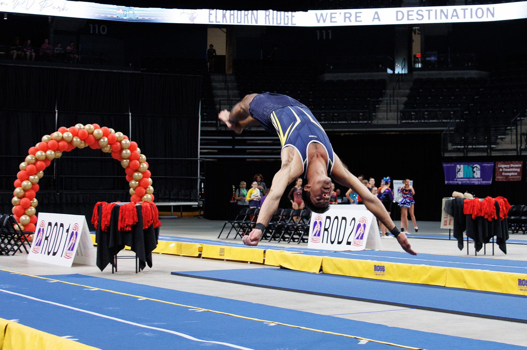A male gymnast performing a tumbling pass on a blue gymnastics floor mat with yellow borders. He is captured mid-flip in an inverted position wearing a dark blue leotard. The scene is set in a large indoor arena with empty stadium seating visible in the background. Red and white balloon decorations and competition signage are positioned around the floor area.